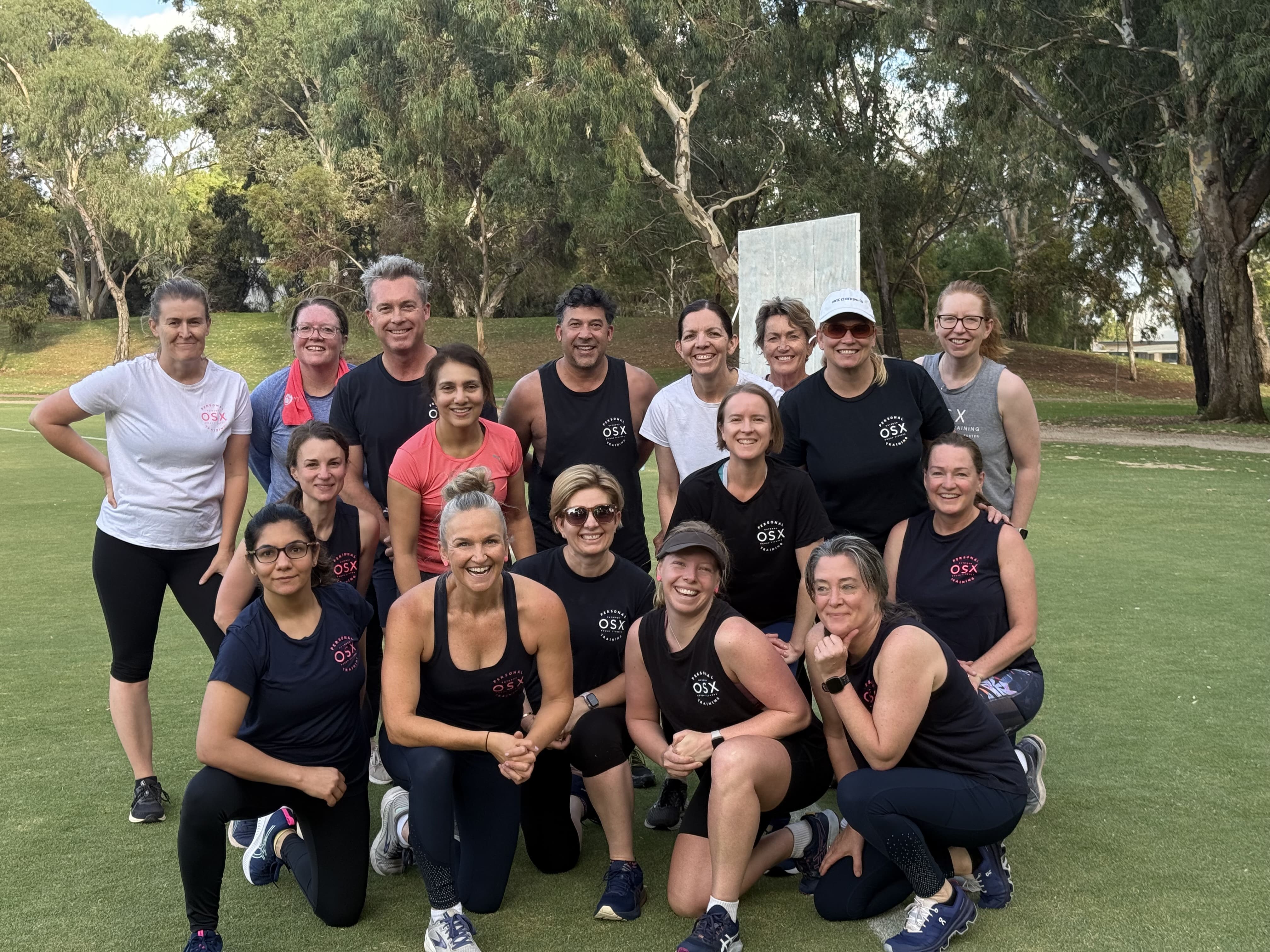 A smiling group of OSX Personal Training members and fitness trainers posing together on a grassy field at an outdoor bootcamp class in Adelaide.