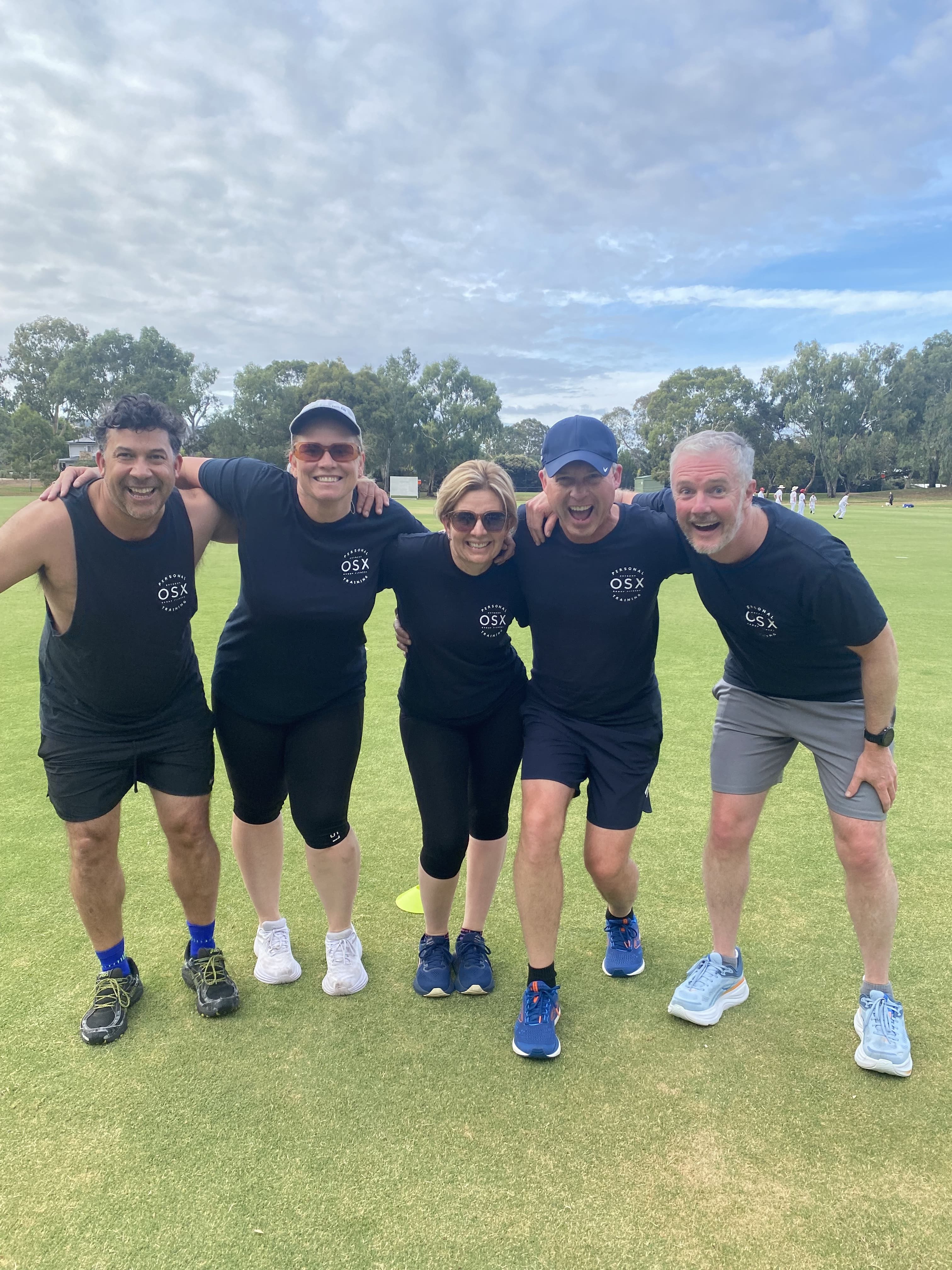 Five smiling OSX Personal Training members standing with arms around each other on an outdoor sports oval in Adelaide